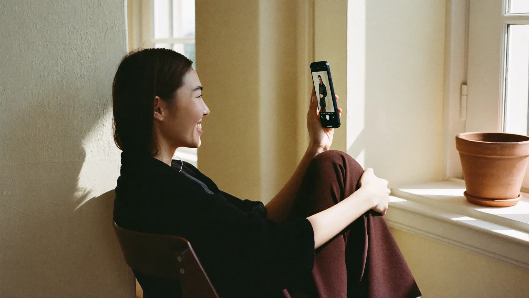A woman sitting by a window in daylight, on a video call, smiling at her phone.