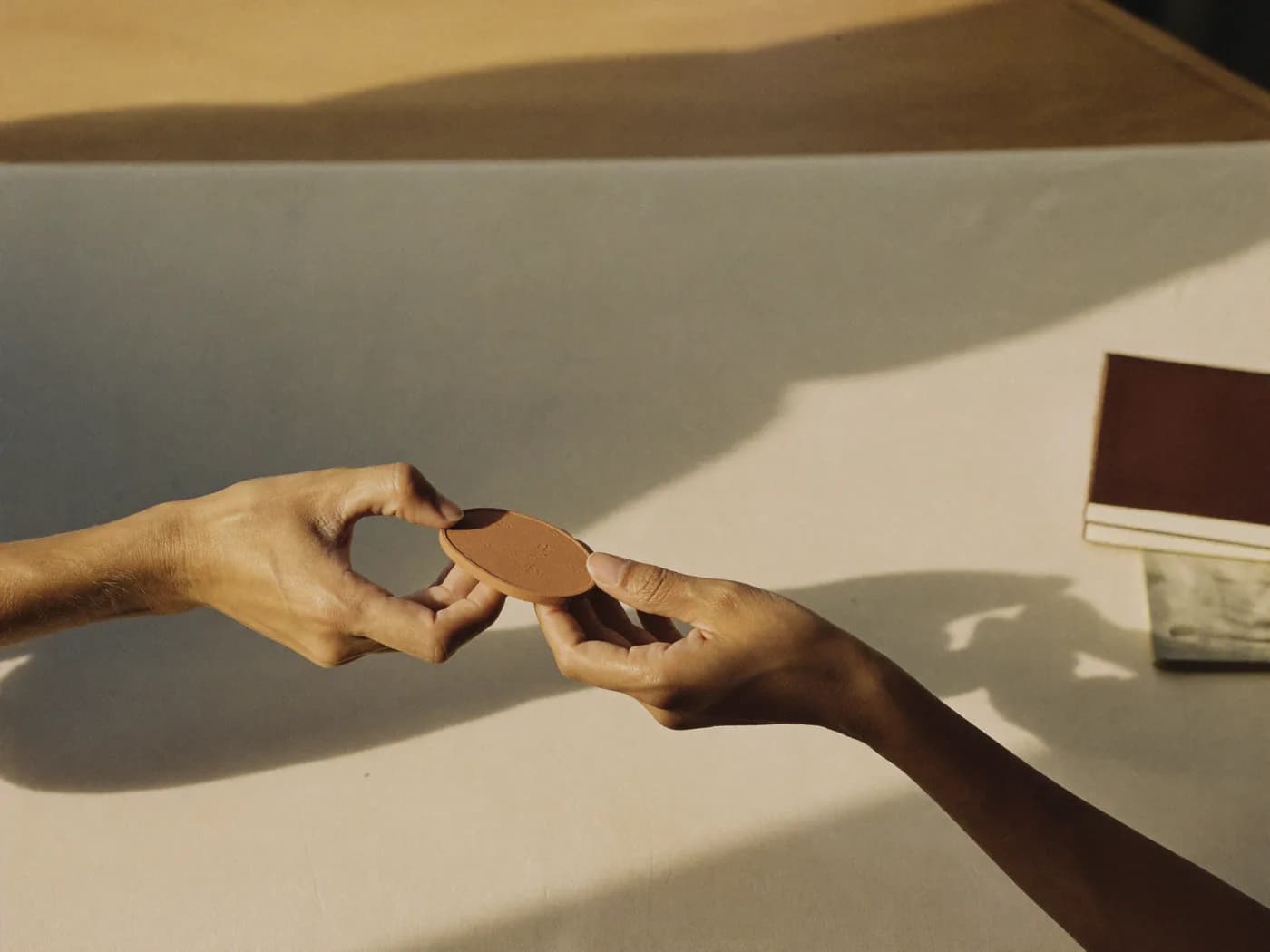 Two hands passing a small terracotta token across a sunlit table.